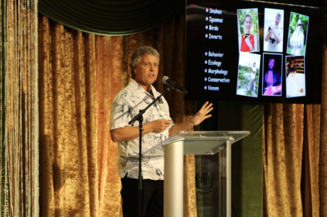 Bill Hayes standin on stage behind a podium with a presentation screen to the side.